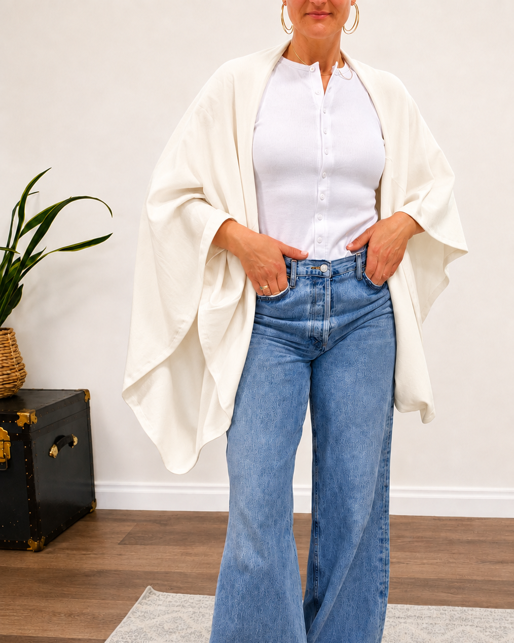 A woman models a white button-down shirt paired with wide-leg blue jeans, a draped cream poncho-style cardigan, and yellow slide sandals in a bright indoor setting.