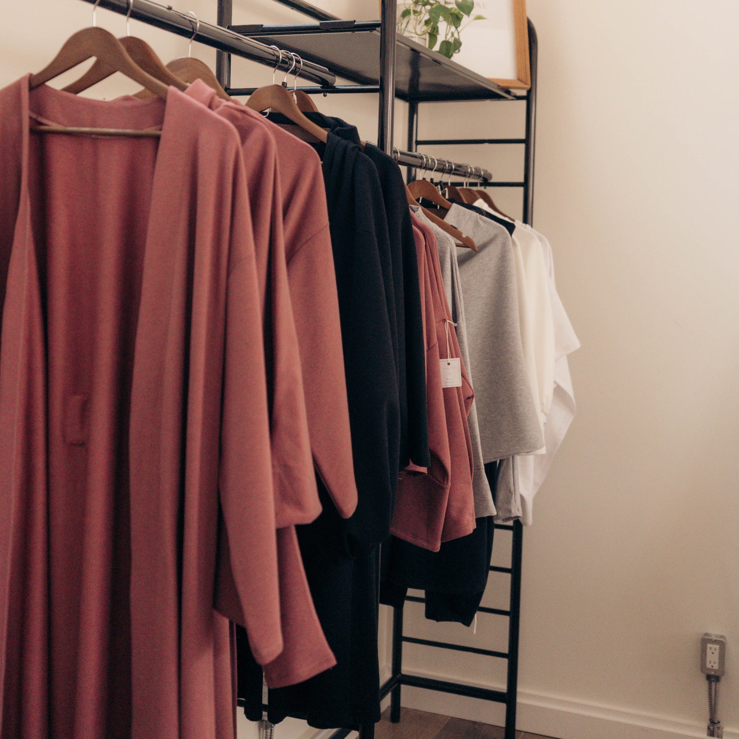 A black metal clothing rack in a minimalist room, holding a dusty rose cardigan, a black top, and other garments, with a small framed print on the top shelf.