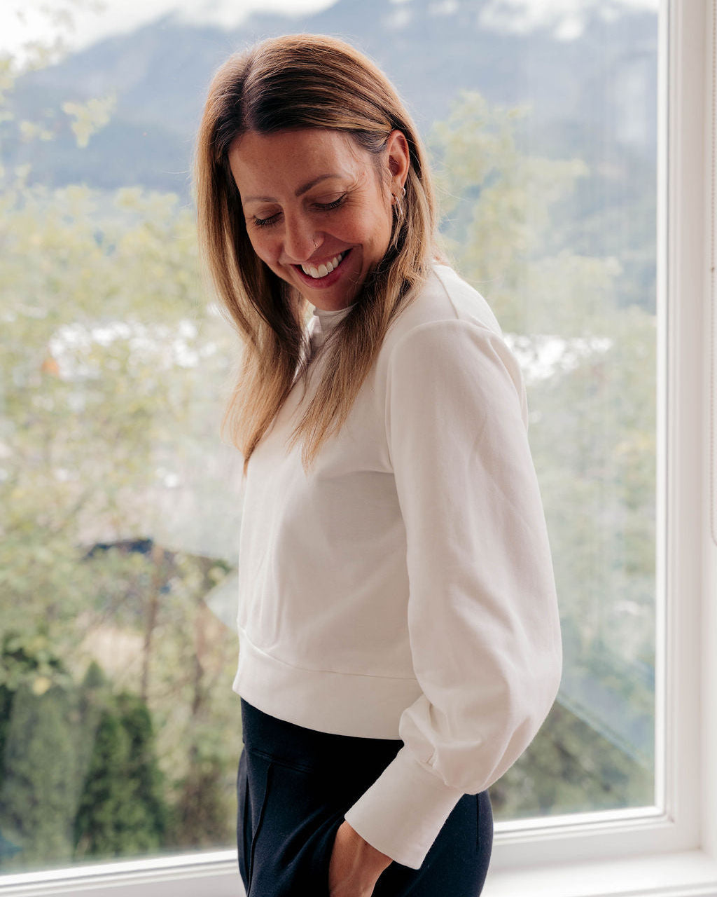 A woman with blonde hair smiling and looking down, wearing a white long-sleeve top and dark pants, standing in front of a large window with a mountain and forest view in the background.