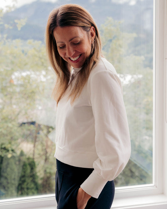 A woman with blonde hair smiling and looking down, wearing a white long-sleeve top and dark pants, standing in front of a large window with a mountain and forest view in the background.