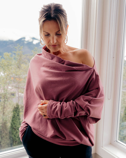 A woman wearing a dusty rose off-the-shoulder long-sleeve top and black pants, standing by a window with a mountain view in the background.