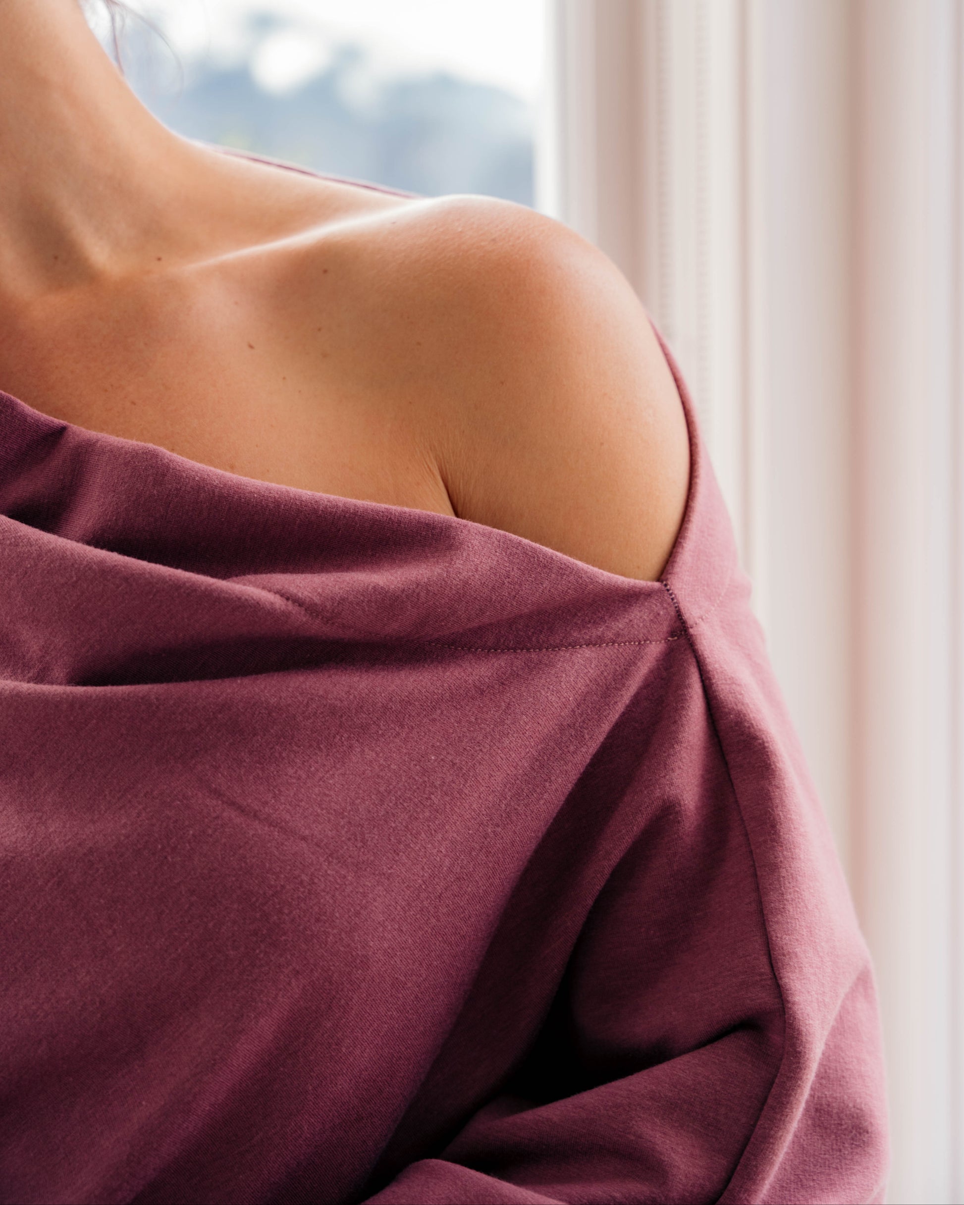 A close-up lifestyle shot of a woman from behind, wearing a dusty rose off-the-shoulder top or wrap, standing near a bright window with soft light filtering in.