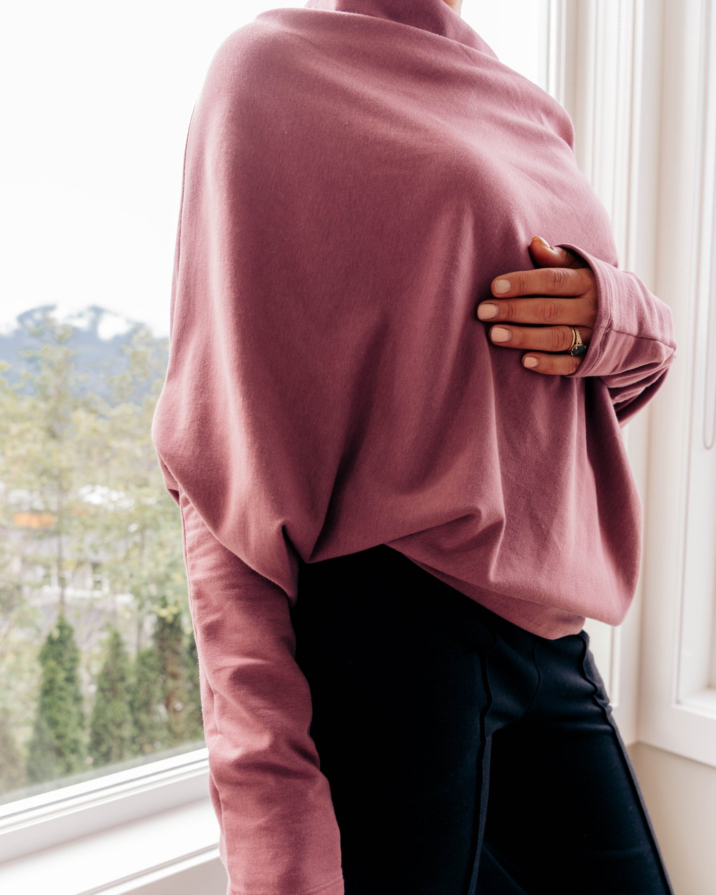 A person wearing a dusty rose long-sleeve knit top and black pants, standing by a bright window.