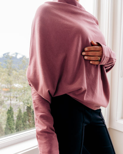 A person wearing a dusty rose long-sleeve knit top and black pants, standing by a bright window.