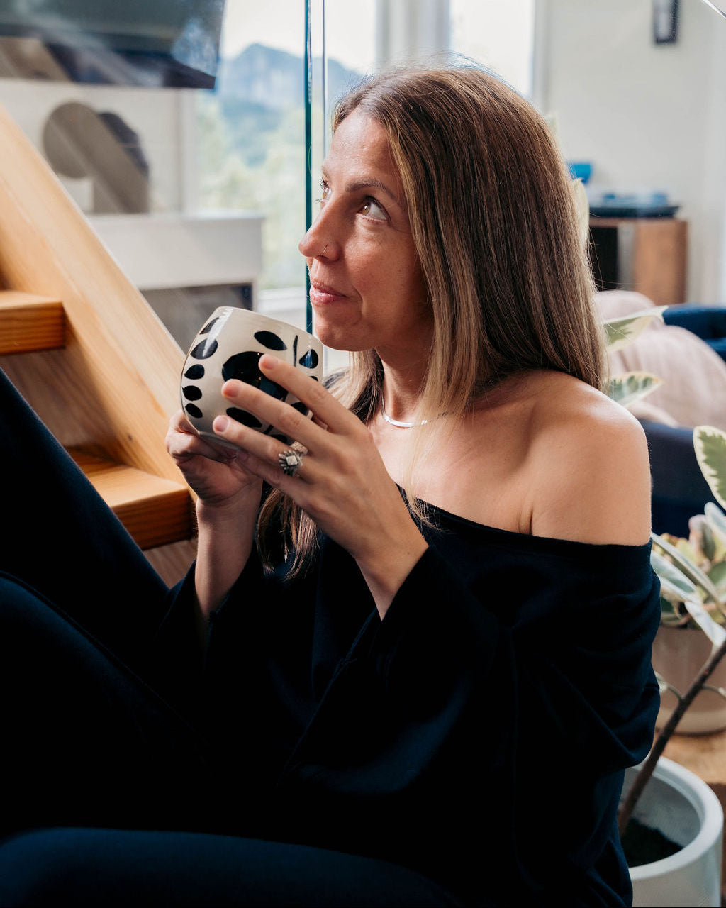 A woman with shoulder-length brown hair sits indoors near a glass wall, holding a black-and-white patterned ceramic mug. She is wearing a dark off-the-shoulder top and black pants in a modern interior setting with natural light.
