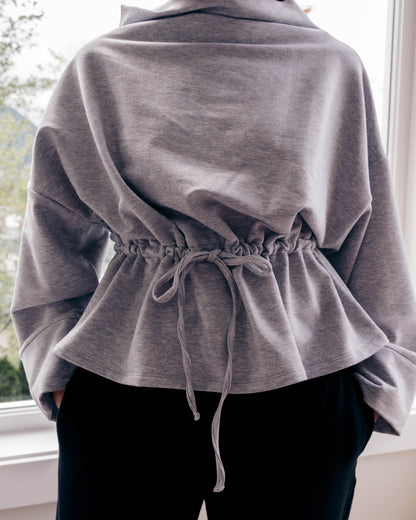 A person wearing a heather gray cropped hoodie with a drawstring waist and black pants, standing indoors near a window.