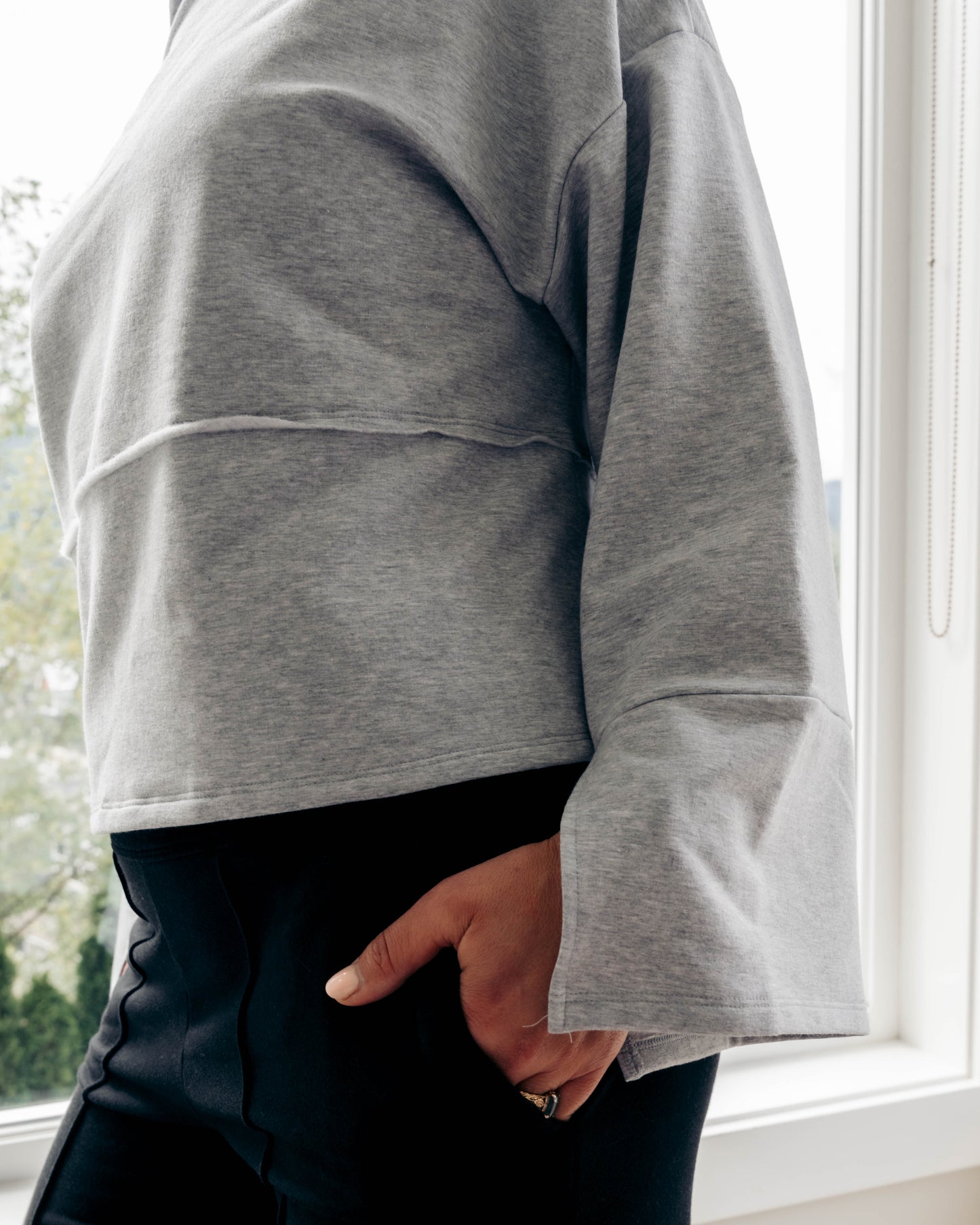 A person wearing a light gray cropped sweatshirt with a layered hem and flared cuffs, paired with black pants, standing by a bright window.