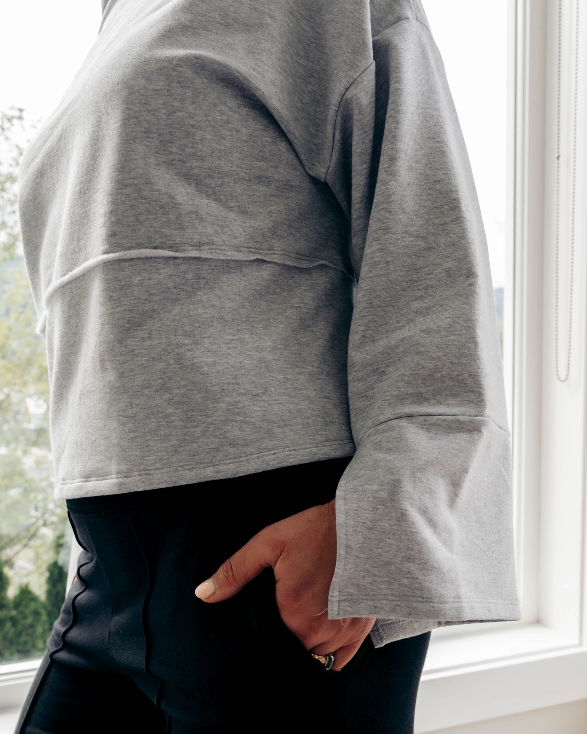 A person wearing a light gray cropped sweatshirt with a layered hem and flared cuffs, paired with black pants, standing by a bright window.
