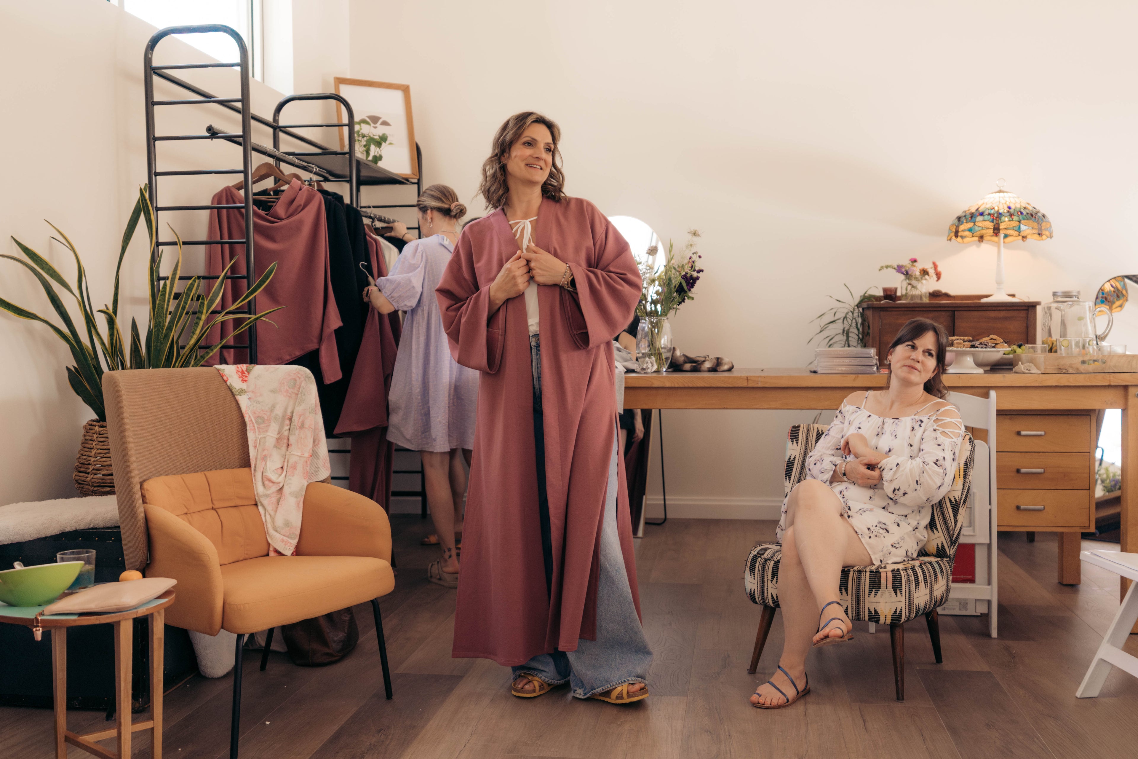 A woman in a dusty rose robe stands in a bright, styled room, while another woman sits in a striped armchair nearby. A clothing rack with more garments is visible in the background.