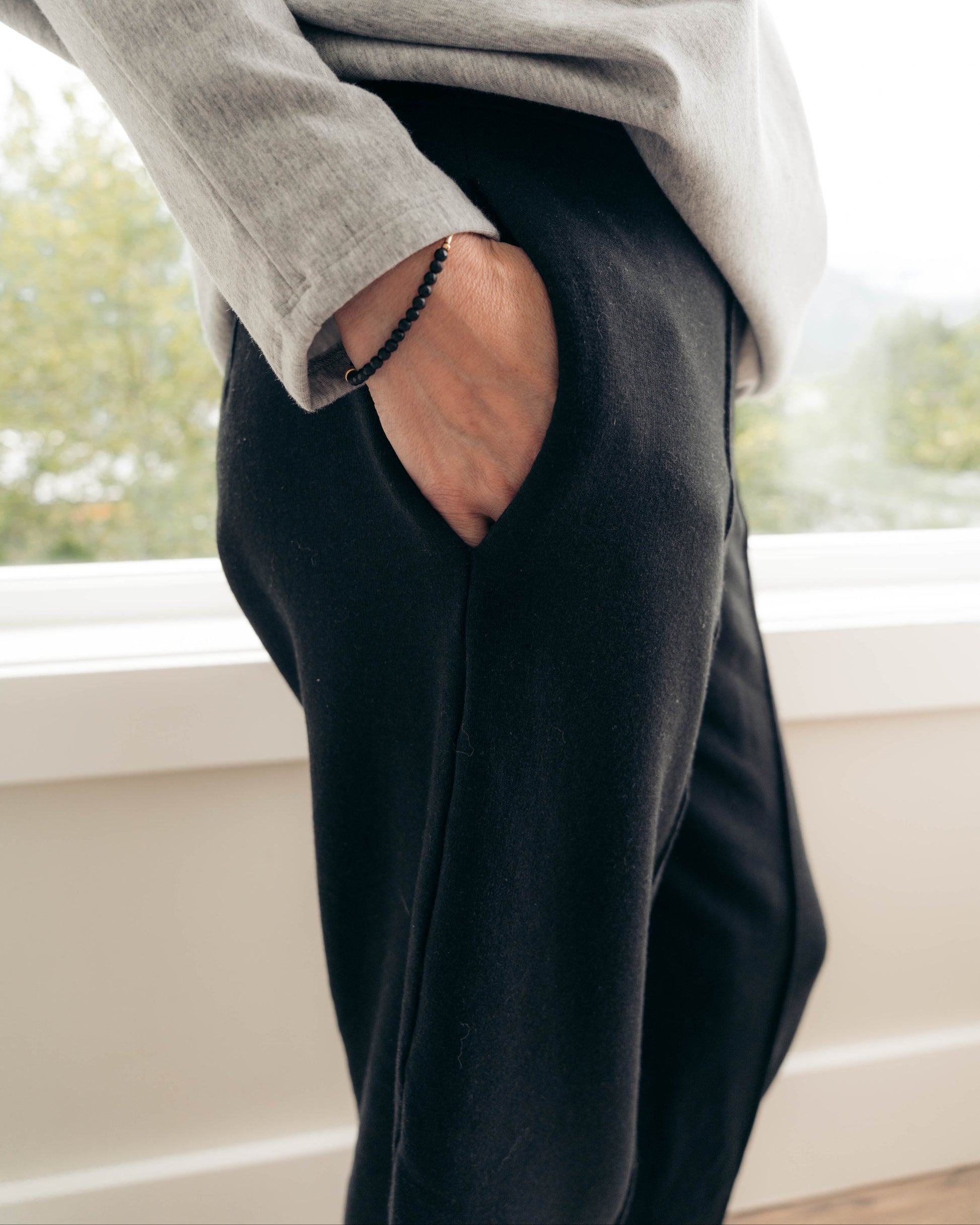 A person wearing black wide-leg lounge pants and a light gray long-sleeve top, standing by a window with their hand in the pocket of the pants.