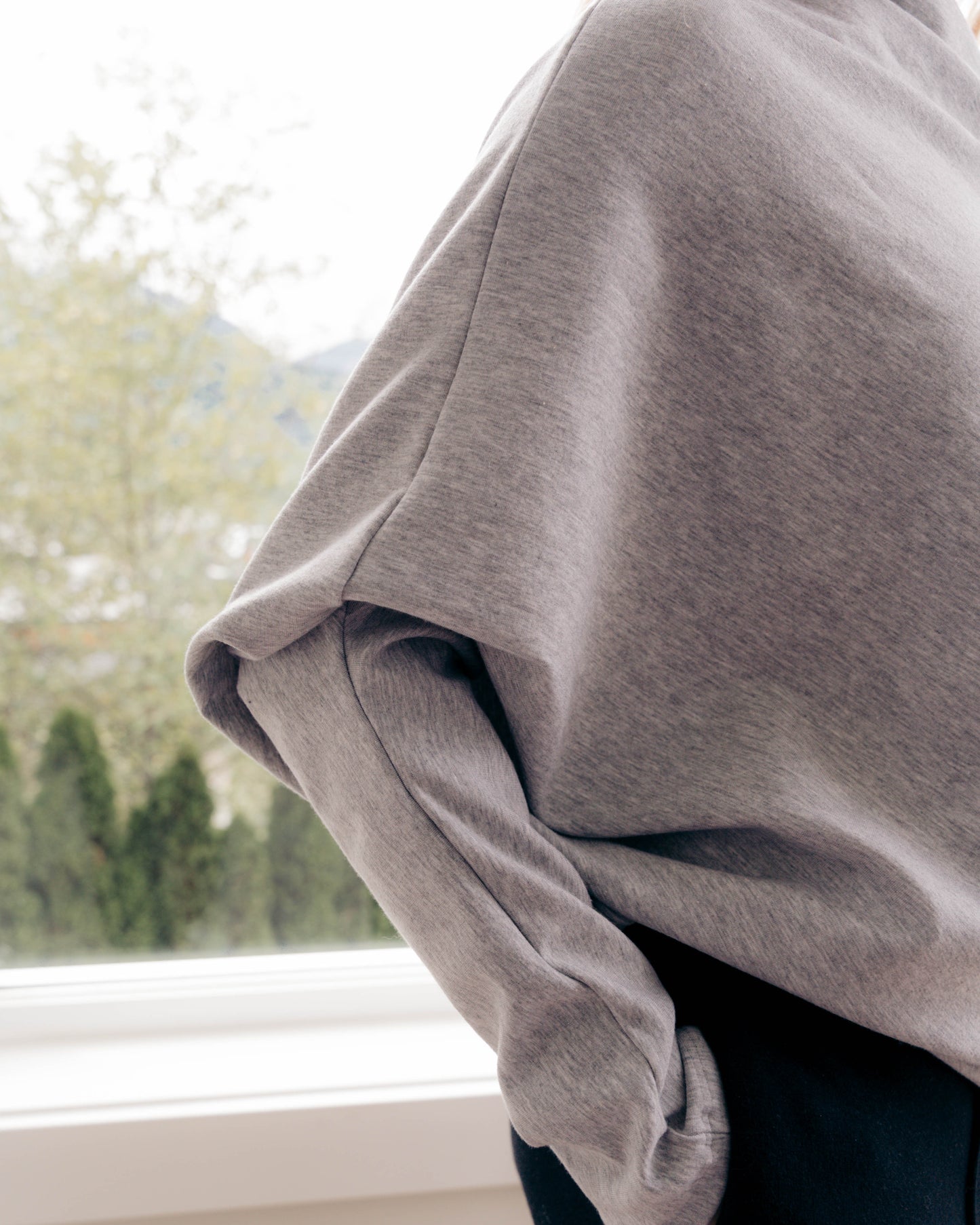 A close-up lifestyle shot of a person wearing an oversized heather gray sweatshirt and dark pants, standing near a window with a blurred outdoor background.