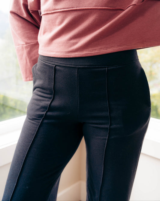 A close-up of a person wearing black high-waisted, structured trousers with vertical seam detailing, paired with a dusty rose long-sleeve top, standing near a window.
