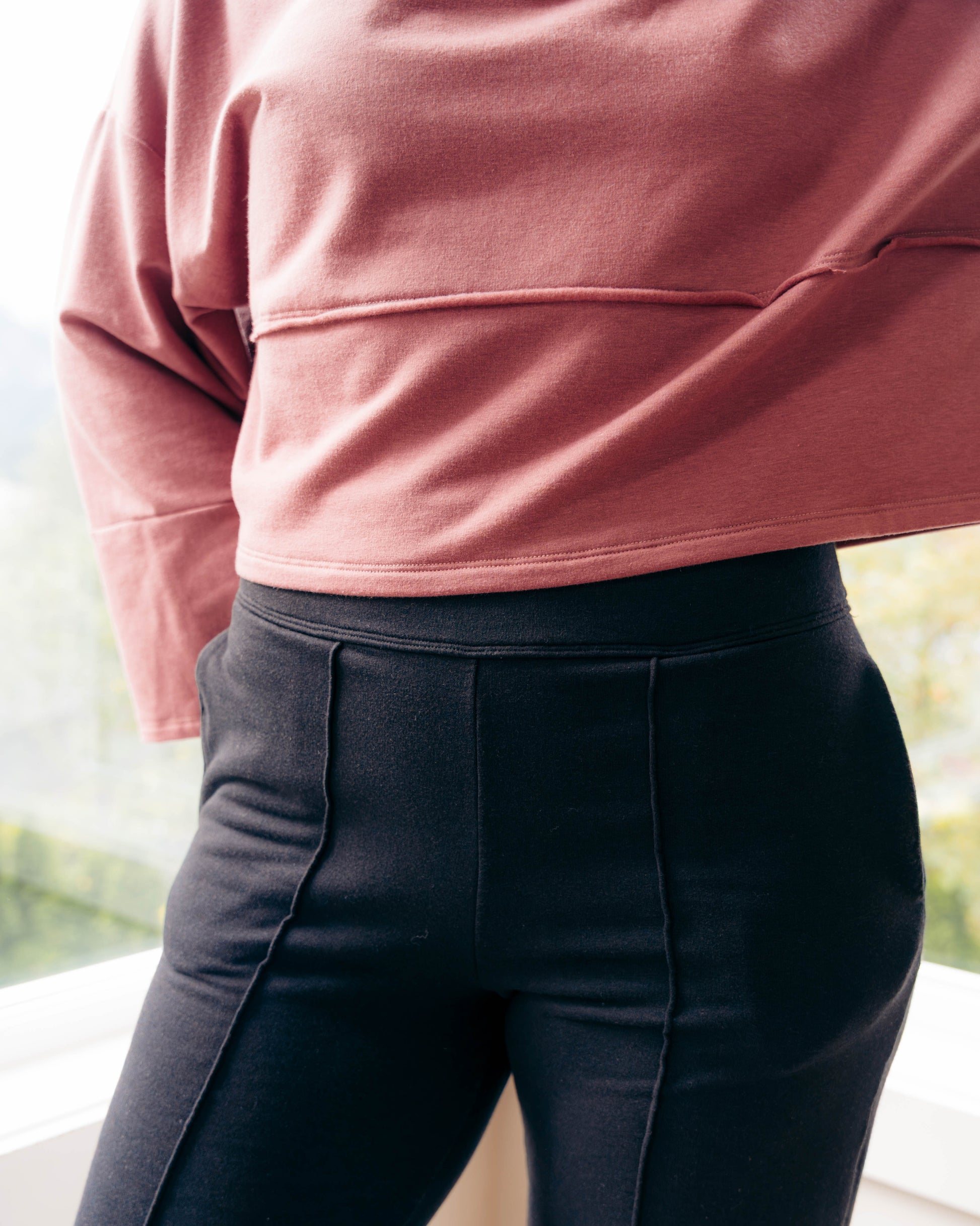 A close-up of a person wearing black high-waisted, structured trousers with vertical seam detailing, paired with a dusty rose long-sleeve top, standing near a window.