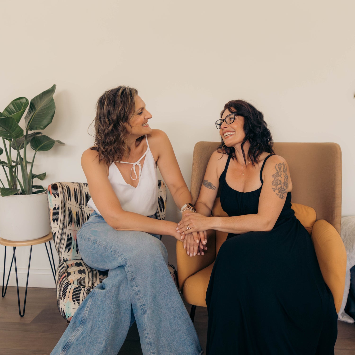 Two women sitting on a couch in a brightly lit room, holding hands and smiling at each other. One wears a white tank top and wide-leg blue jeans, and the other wears a black dress and glasses.