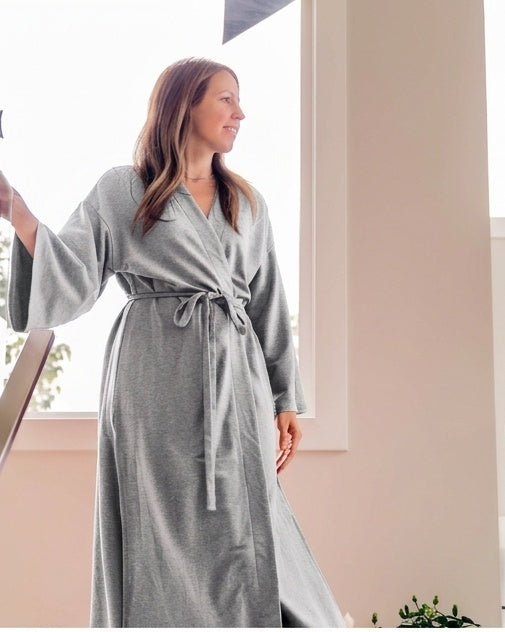 A woman wearing a long gray wrap robe with a tie waist, standing indoors near a bright window.