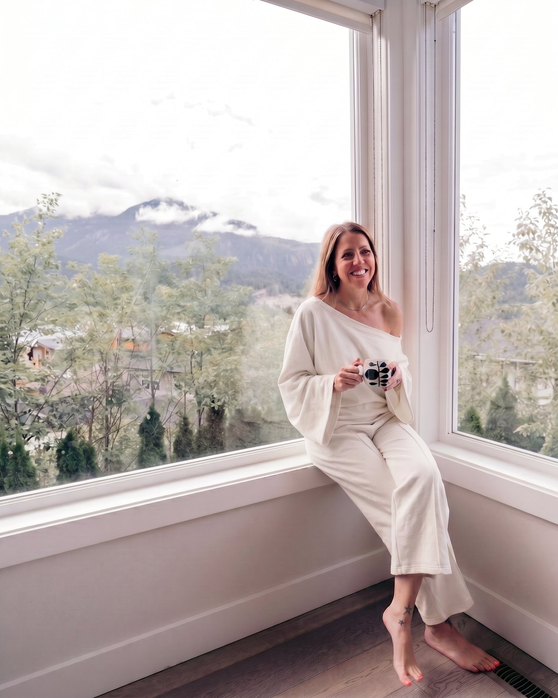 A woman wearing a cream off-the-shoulder long-sleeve jumpsuit sits barefoot on a window sill, holding a camera, with a mountain landscape visible outside.