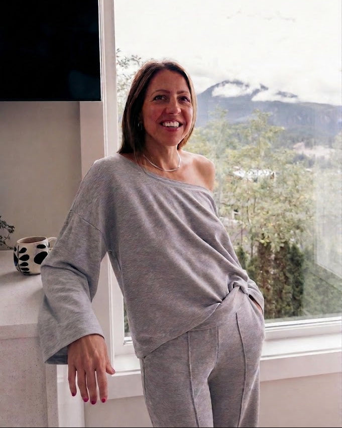 A woman wearing a light gray heathered lounge set consisting of an asymmetrical one-shoulder top and matching pants, standing by a large window with a mountain view.
