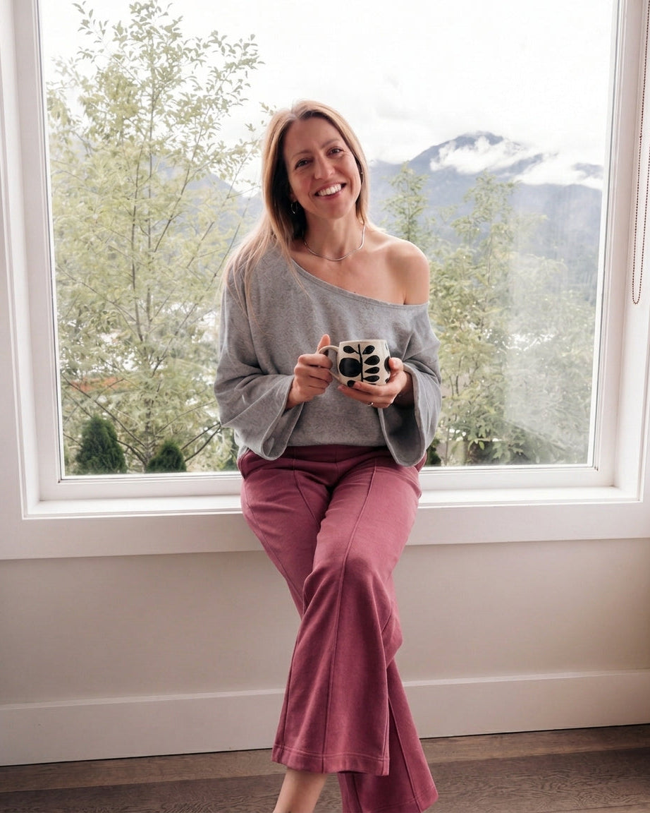 A woman sitting by a large window wearing a gray off-the-shoulder knit sweater and dusty rose flare pants, holding a small camera. The background shows a bright outdoor view of trees and mountains.