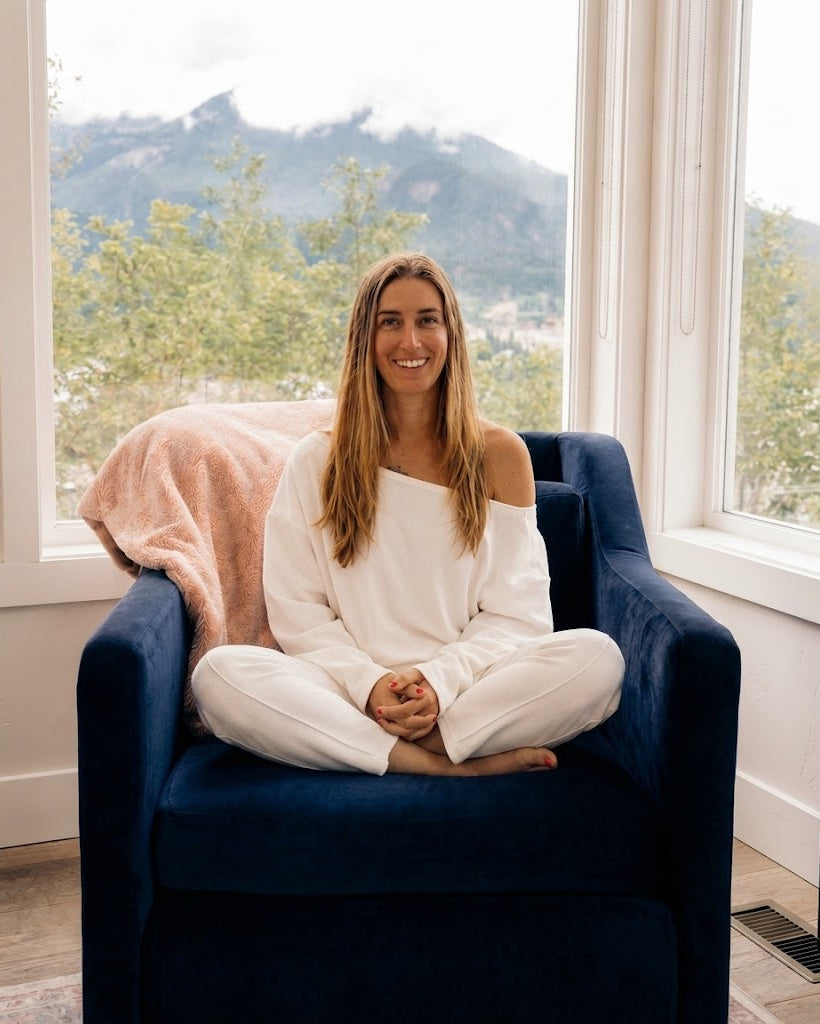 A woman with long light brown hair sits cross-legged in a navy blue armchair in front of large windows with a mountain view. She is wearing a white off-the-shoulder long-sleeve top and matching white pants.
