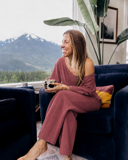 A woman sits barefoot on a dark blue velvet armchair in a living room, wearing a dusty rose off-the-shoulder long-sleeve jumpsuit. She holds a mug, with a mountain landscape visible through the window behind her.