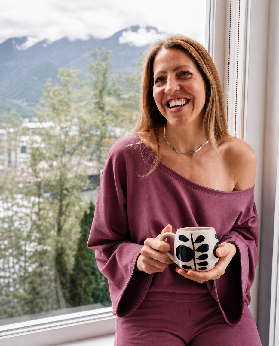 A woman smiling by a window, wearing a matching off-the-shoulder mauve lounge set and holding a white mug with black abstract leaf-like spots. Mountains and trees are visible outside the window.