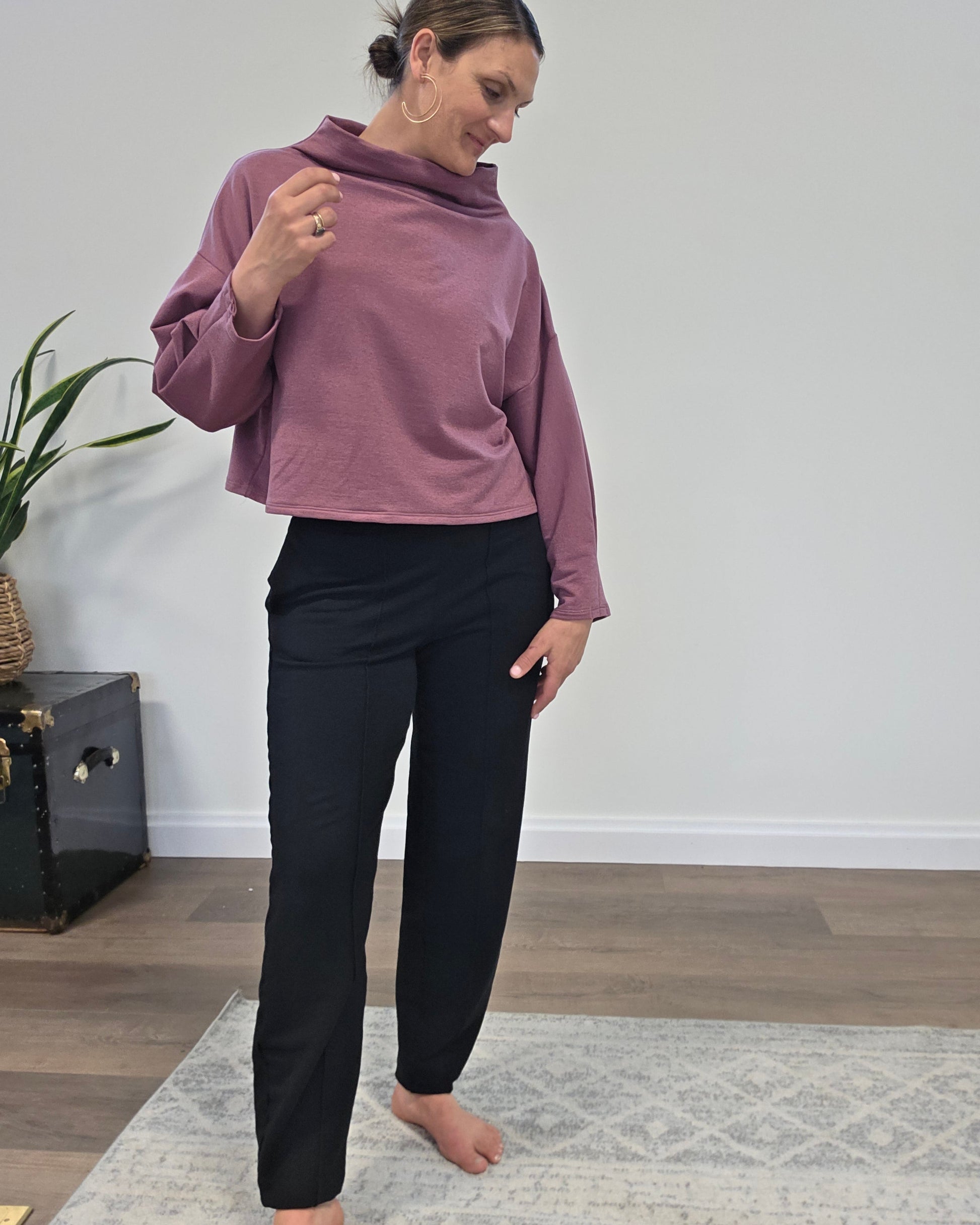 A woman modeling a dusty mauve cropped long-sleeve cowl-neck top and black pants, standing barefoot on a light patterned rug in a minimalist room with wood floors and a white wall.
