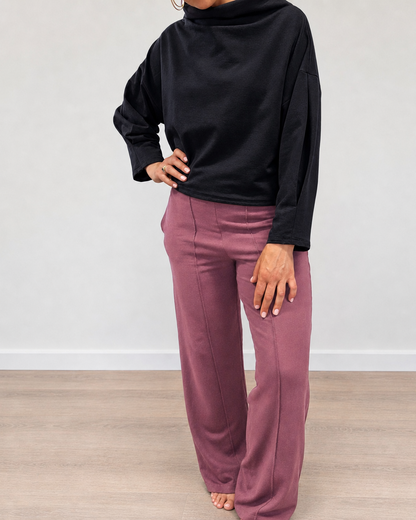 A person wearing a black long-sleeve mock-neck top and dusty rose wide-leg pants, standing barefoot on a light wood floor against a plain light gray wall.
