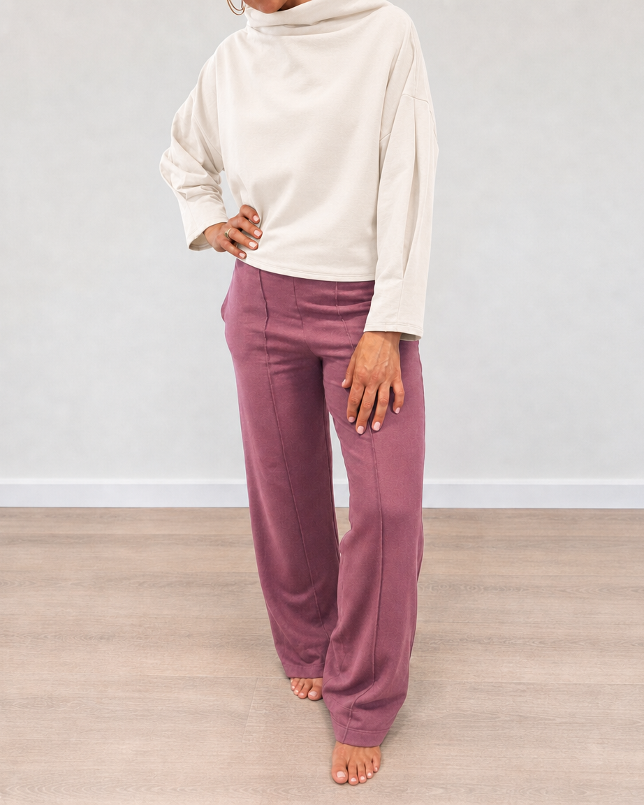 A person wearing a cream mock-neck long-sleeve top and dusty rose wide-leg pants, standing barefoot on a light wood floor against a neutral gray wall.
