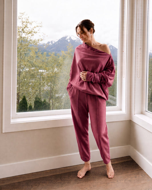 A woman stands barefoot in a room by large windows, wearing a matching dusty rose loungewear set consisting of an asymmetrical long-sleeve top and relaxed pants. Outside the window, mountains and trees are visible.
