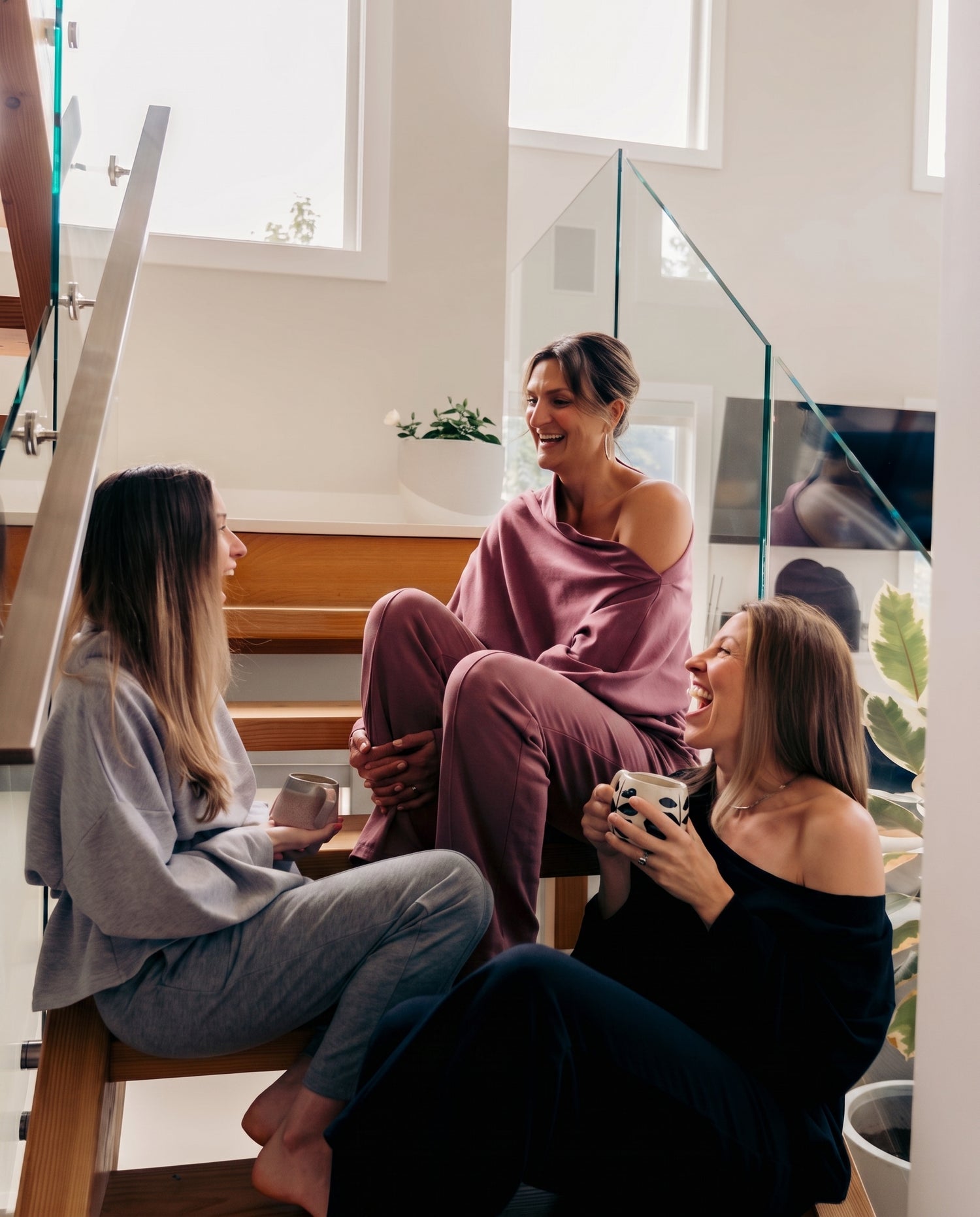 Three women in matching lounge sets sit and relax on wooden stairs in a modern home interior, engaging in conversation and holding mugs.