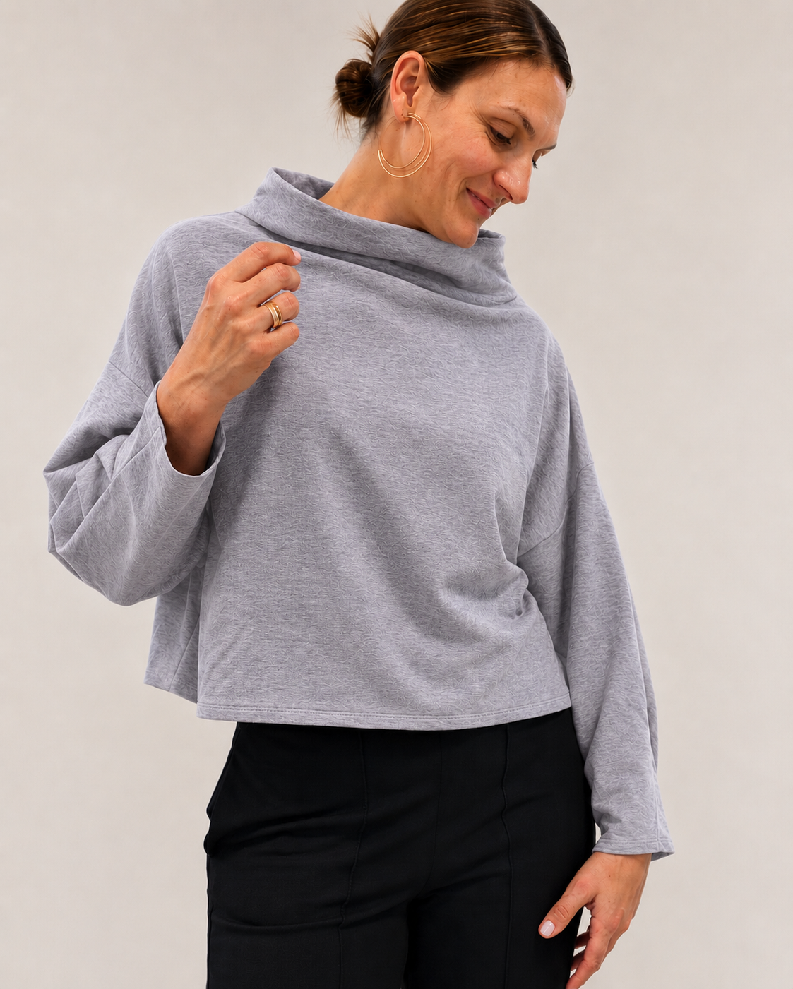 A woman wearing a light gray heathered long-sleeve cowl neck top and black pants, posing against a neutral light gray background.