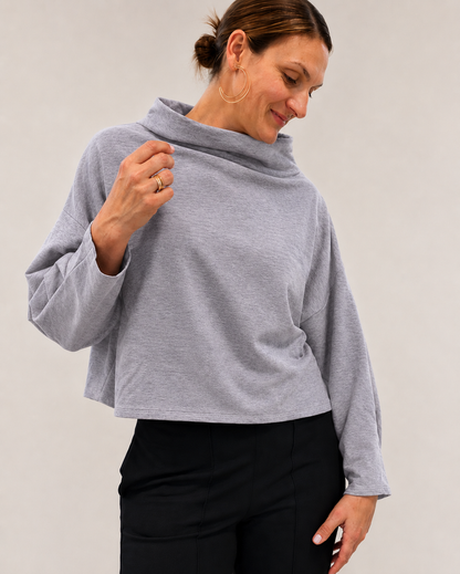 A woman wearing a light gray heathered long-sleeve cowl neck top and black pants, posing against a neutral light gray background.