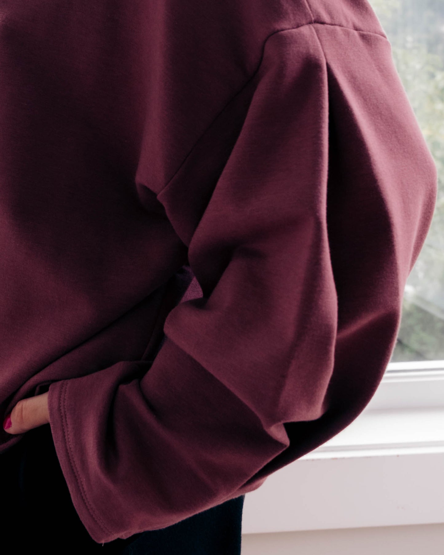 A close-up of a person wearing an oversized long-sleeve sweatshirt in a deep burgundy or maroon shade, standing near a white window frame.