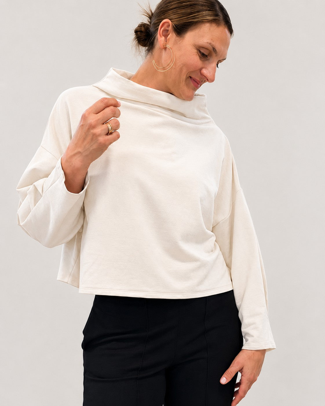 A woman wearing a cream-colored, long-sleeve cropped top with a high folded collar, paired with black pants, against a plain light gray background.