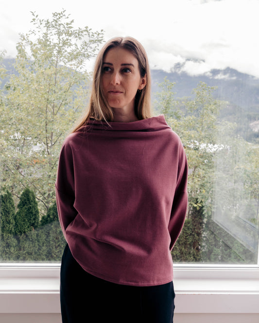 A woman wearing a dusty rose cowl-neck sweatshirt and black pants, standing in front of a large window with a view of trees and mountains in the background.