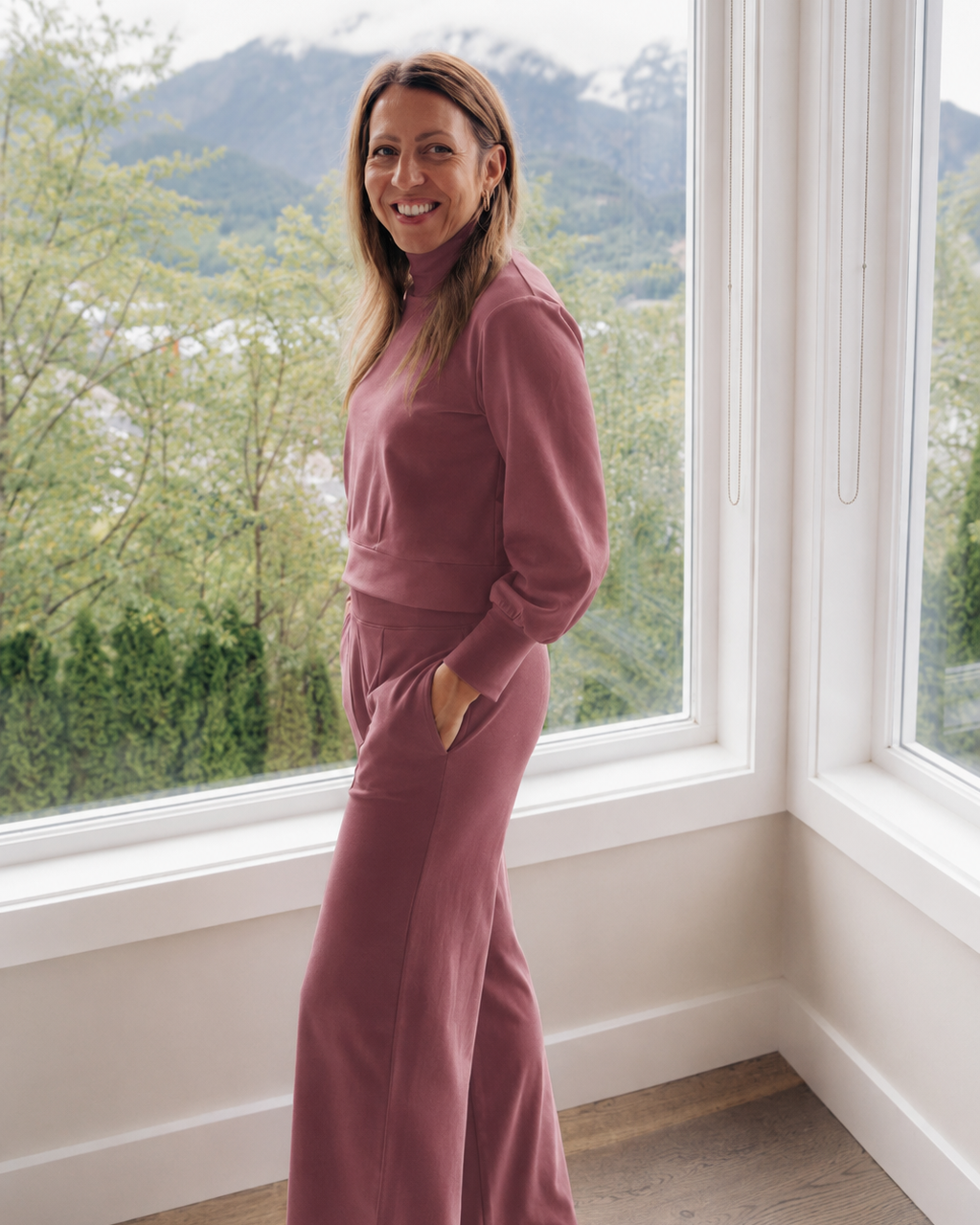 A woman wearing a matching dusty rose two-piece lounge set stands by a large window with a mountain and forest view in the background.