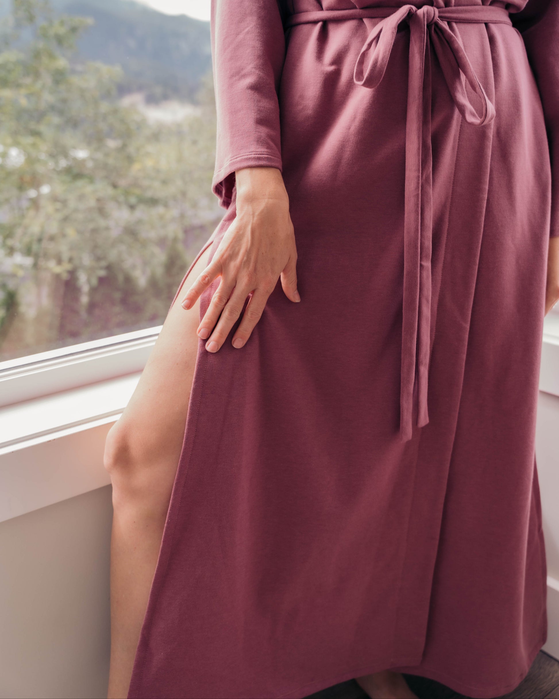 A woman wearing a long-sleeve burnt rose wrap robe with a tie waist belt, standing by a window with a scenic view in the background.