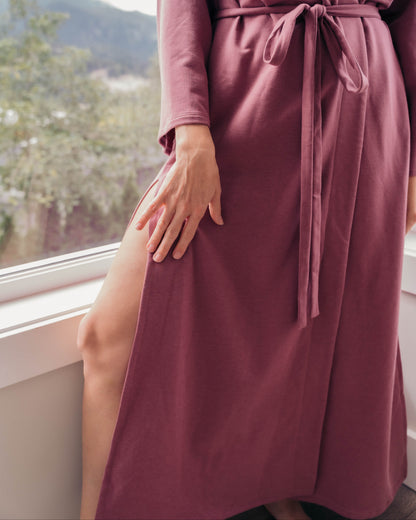 A woman wearing a long-sleeve burnt rose wrap robe with a tie waist belt, standing by a window with a scenic view in the background.