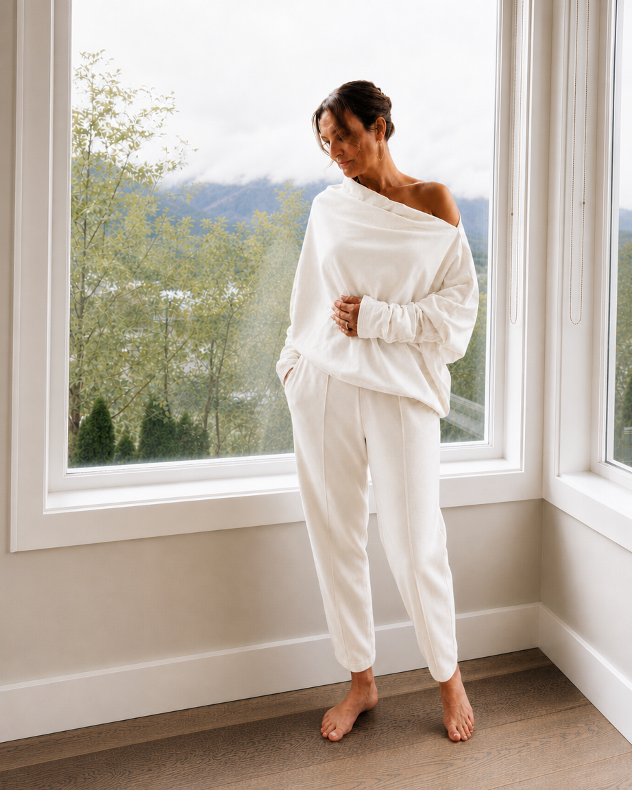 A woman stands barefoot in a bright room by a large window, wearing a loose off-white long-sleeve top and matching off-white pants. The background shows a soft-focus outdoor view of trees and hills.