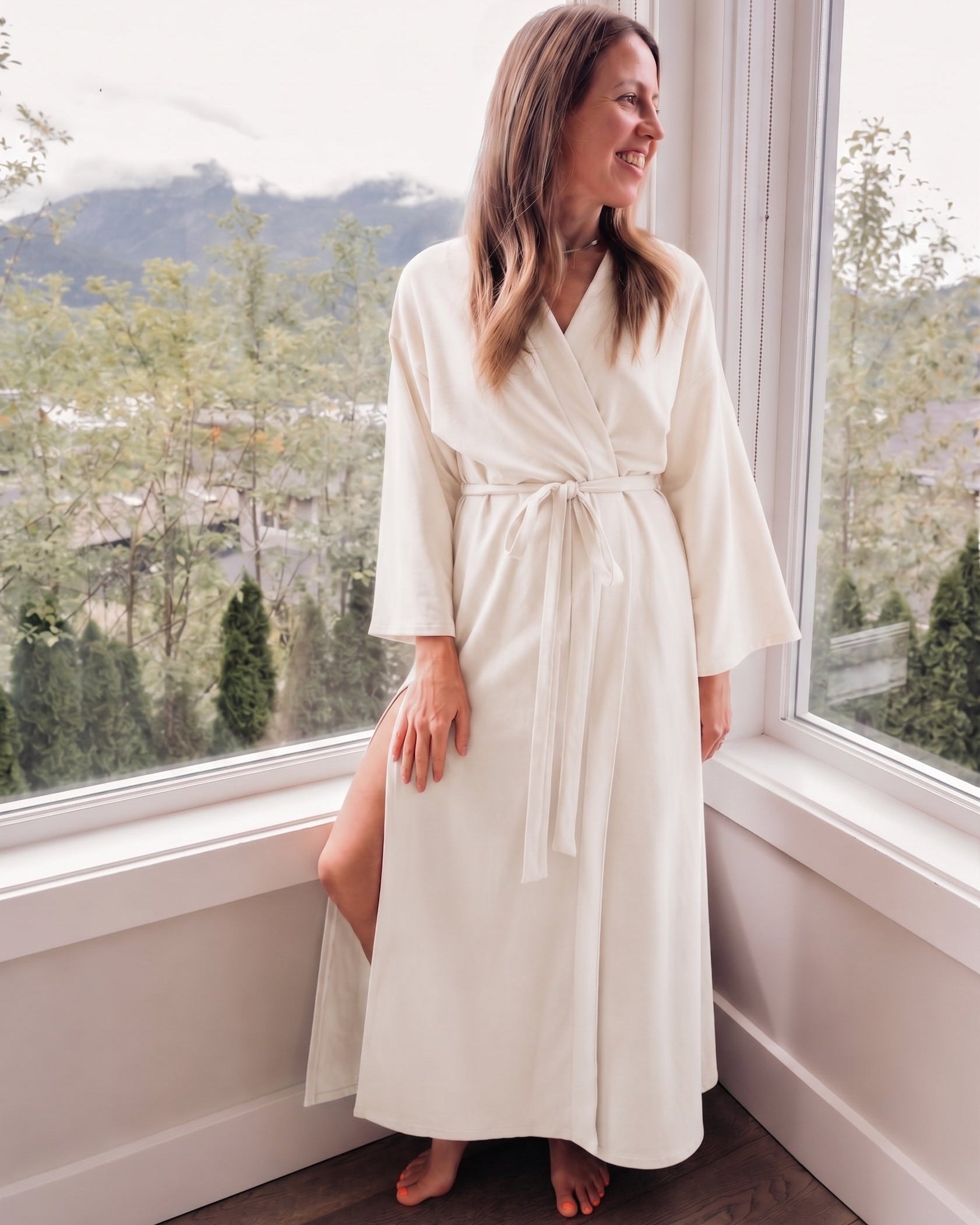 A woman wearing a long ivory-colored robe stands barefoot by a large window with a mountain and tree view outside.