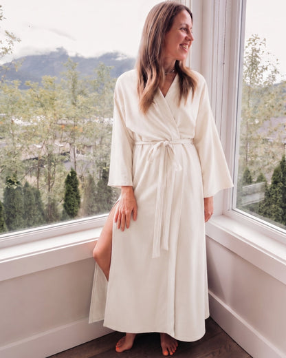 A woman wearing a long ivory-colored robe stands barefoot by a large window with a mountain and tree view outside.