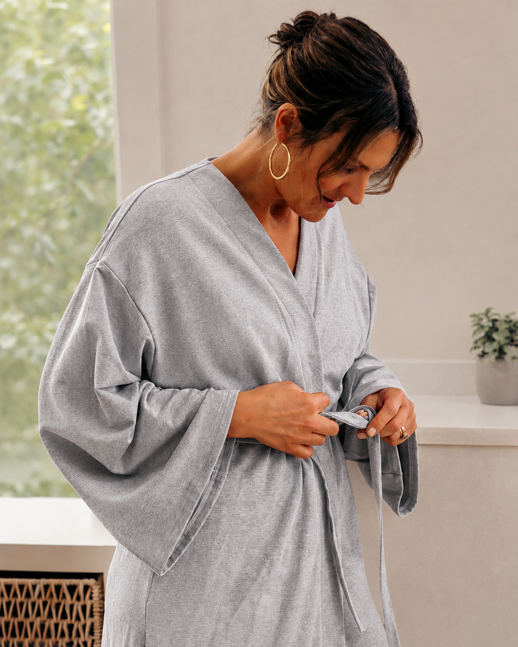 A woman wearing a light gray oversized robe with wide sleeves and a tie belt, standing indoors near a window and counter.