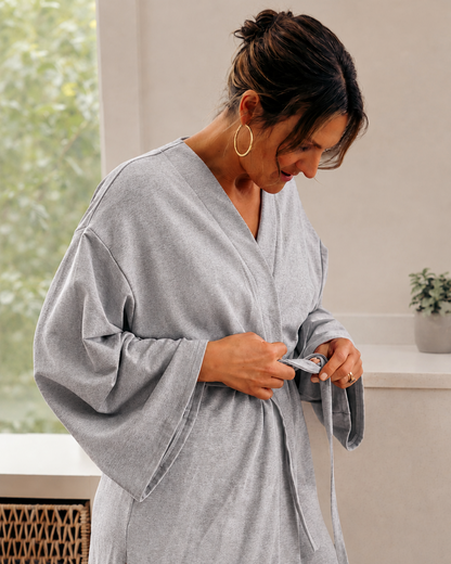 A woman wearing a light gray oversized robe with wide sleeves and a tie belt, standing indoors near a window and counter.