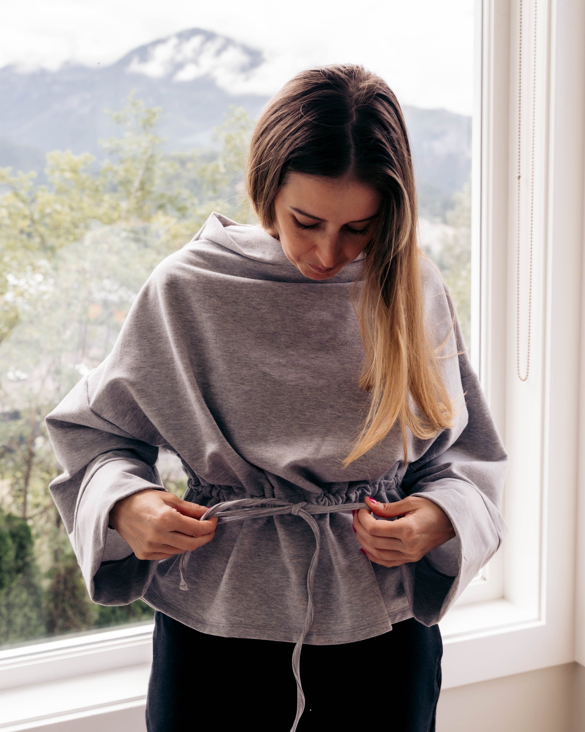 A woman wearing a gray wrap poncho-style top with a drawstring waist, standing by a window with a mountain landscape visible outside.