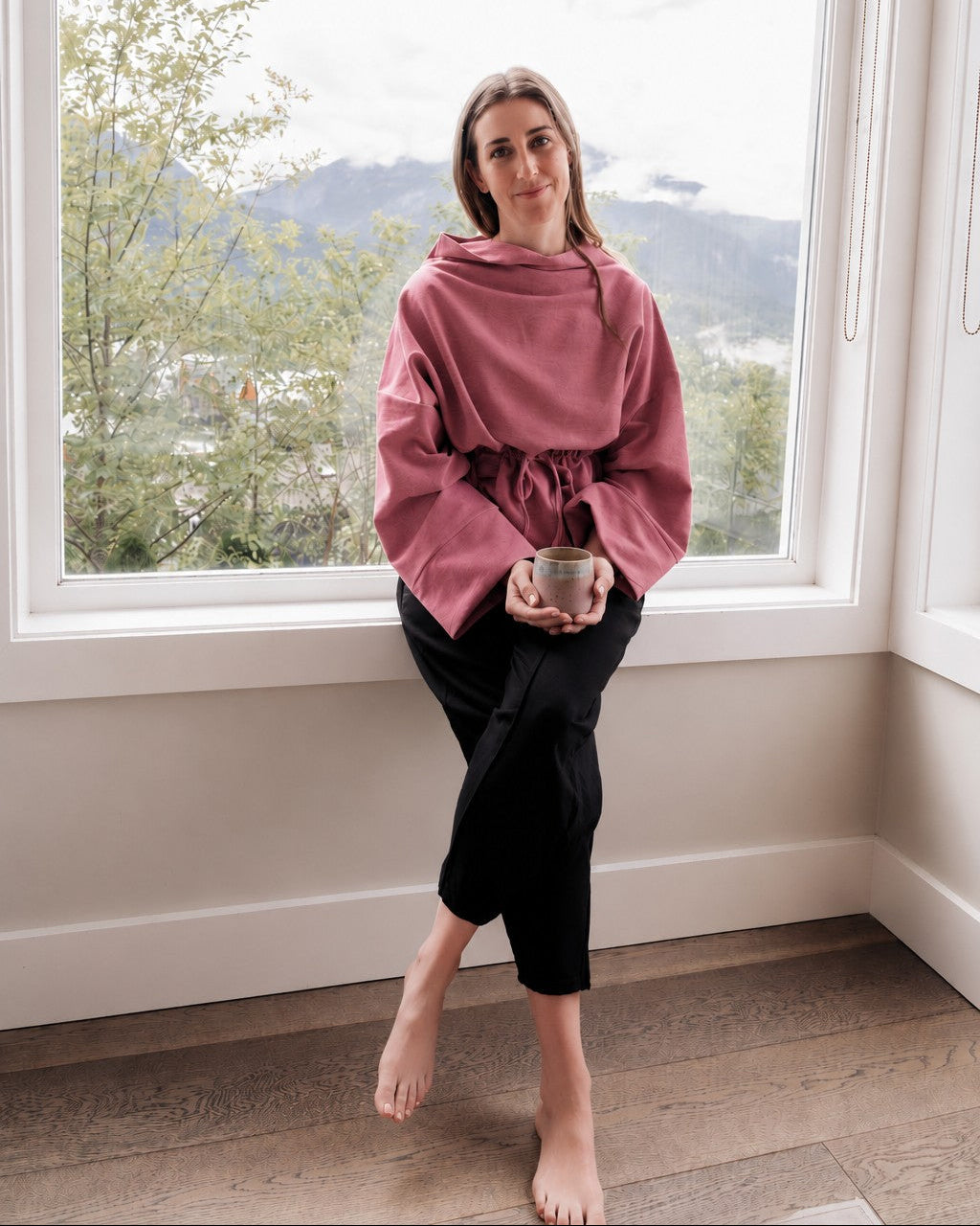 A woman sits barefoot on a window sill in a room with a mountain view, wearing a dusty rose oversized hoodie with a drawstring waist and black cropped pants, holding a small bowl.