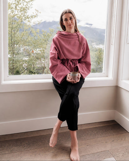 A woman sits barefoot on a window sill in a room with a mountain view, wearing a dusty rose oversized hoodie with a drawstring waist and black cropped pants, holding a small bowl.