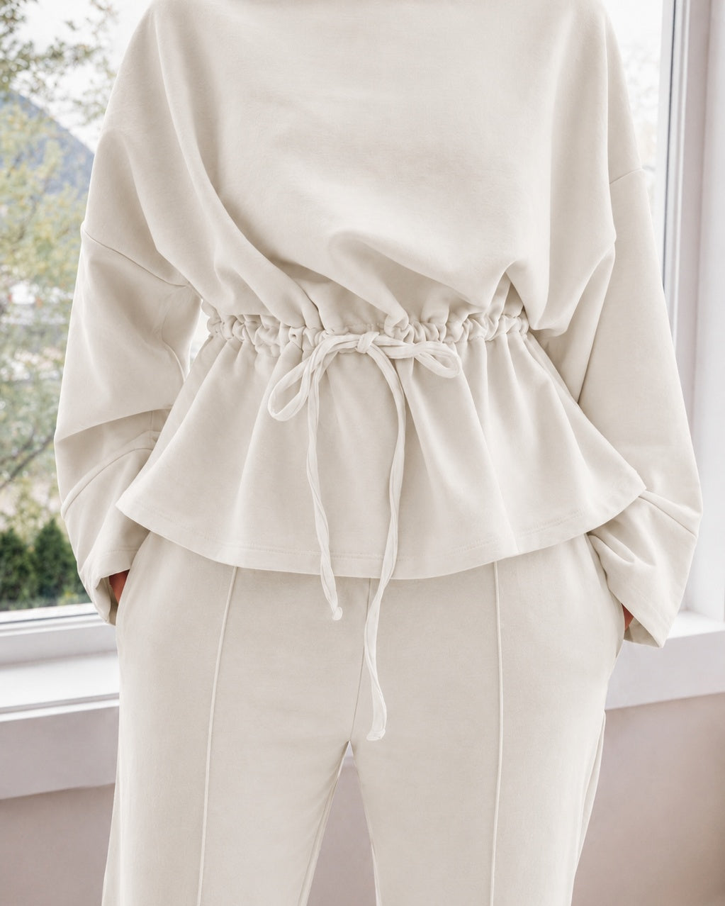 A person wearing an ivory-colored two-piece loungewear set consisting of a long-sleeve top with a drawstring waist and matching drawstring pants, standing in front of a window.