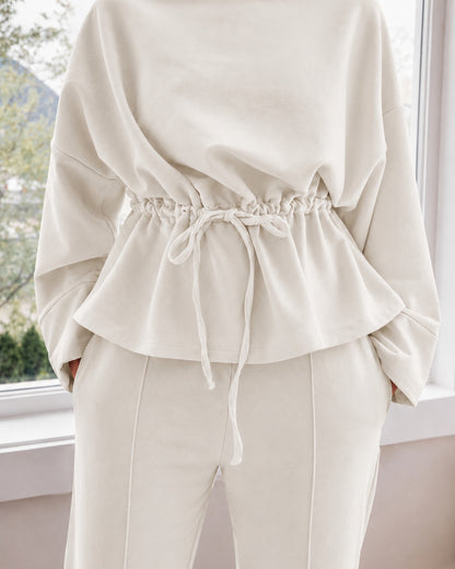 A person wearing an ivory-colored two-piece loungewear set consisting of a long-sleeve top with a drawstring waist and matching drawstring pants, standing in front of a window.