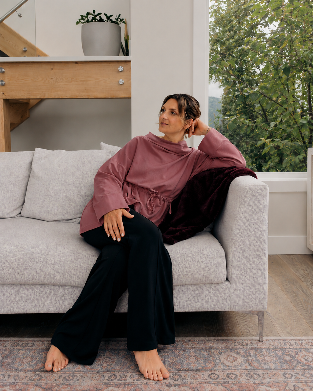 A woman sits barefoot on a light gray sofa in a modern living room, wearing a burnt rose long-sleeve draped top and black wide-leg pants. A dark burgundy velvet throw blanket is draped over the arm of the sofa, and a large window behind her shows green trees outside.