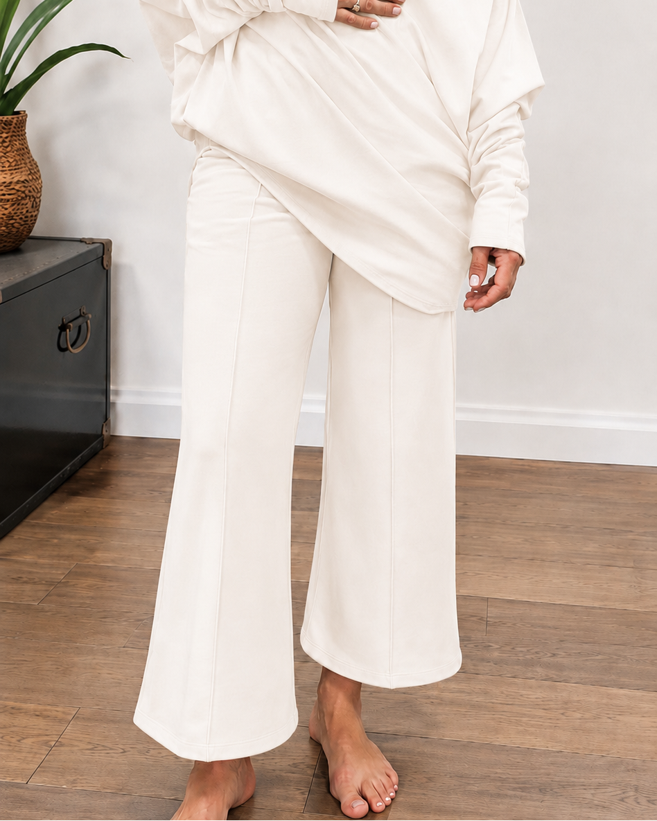 A model wearing a matching off-white two-piece lounge set consisting of a draped long-sleeve top and wide-leg cropped pants, standing on a wooden floor.